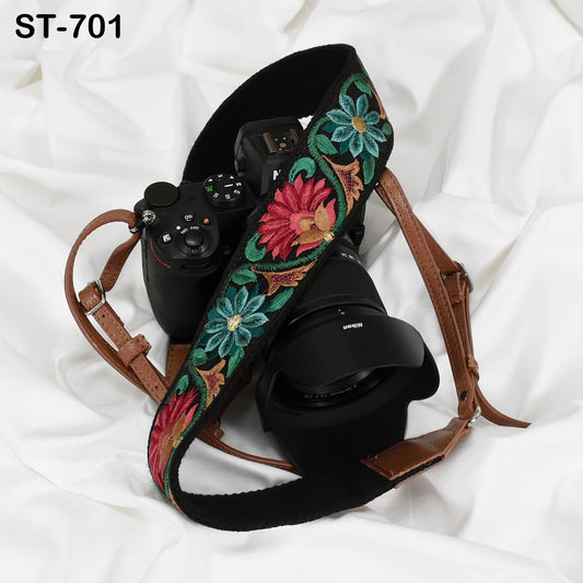 A black camera strap with a floral pattern is draped over a camera lens, resting on a white fabric background.