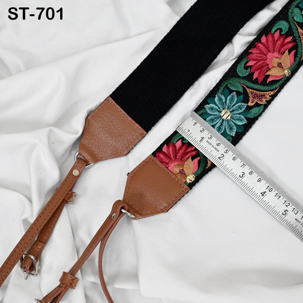 a black and brown camera strap with a floral embroidered pattern, placed on a white fabric surface. A ruler is also visible in the image, indicating the strap's length.