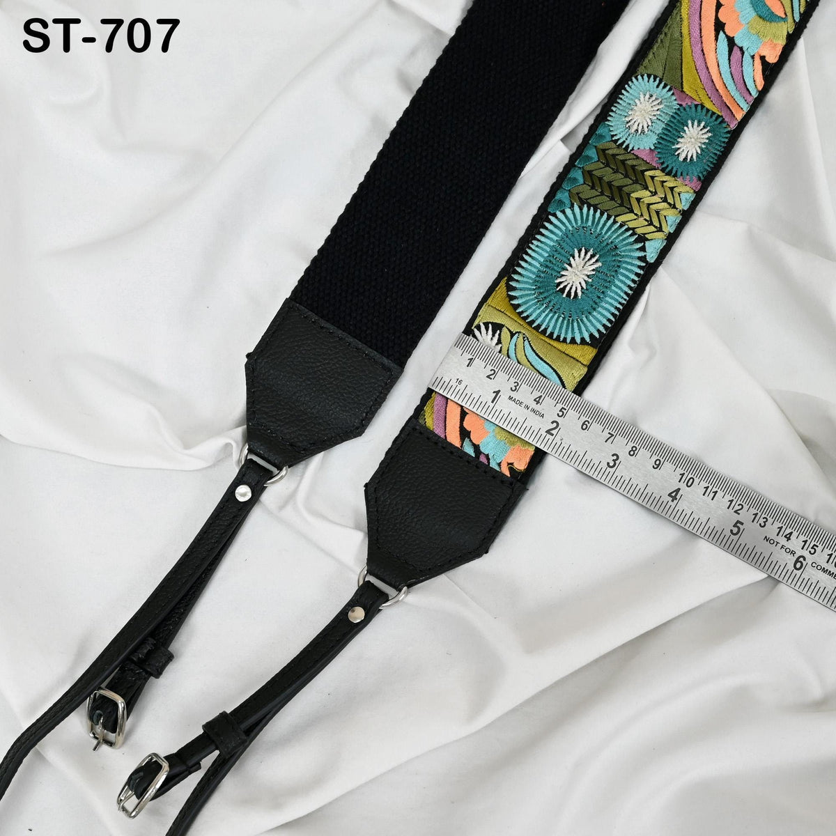 a black camera strap with a colorful floral pattern, placed on a white fabric surface. A ruler is also visible in the image, indicating the strap's length.