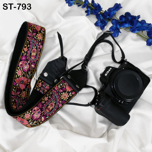 A floral patterned camera strap and a Nikon camera are placed on a white fabric surface, with blue flowers visible in the background.