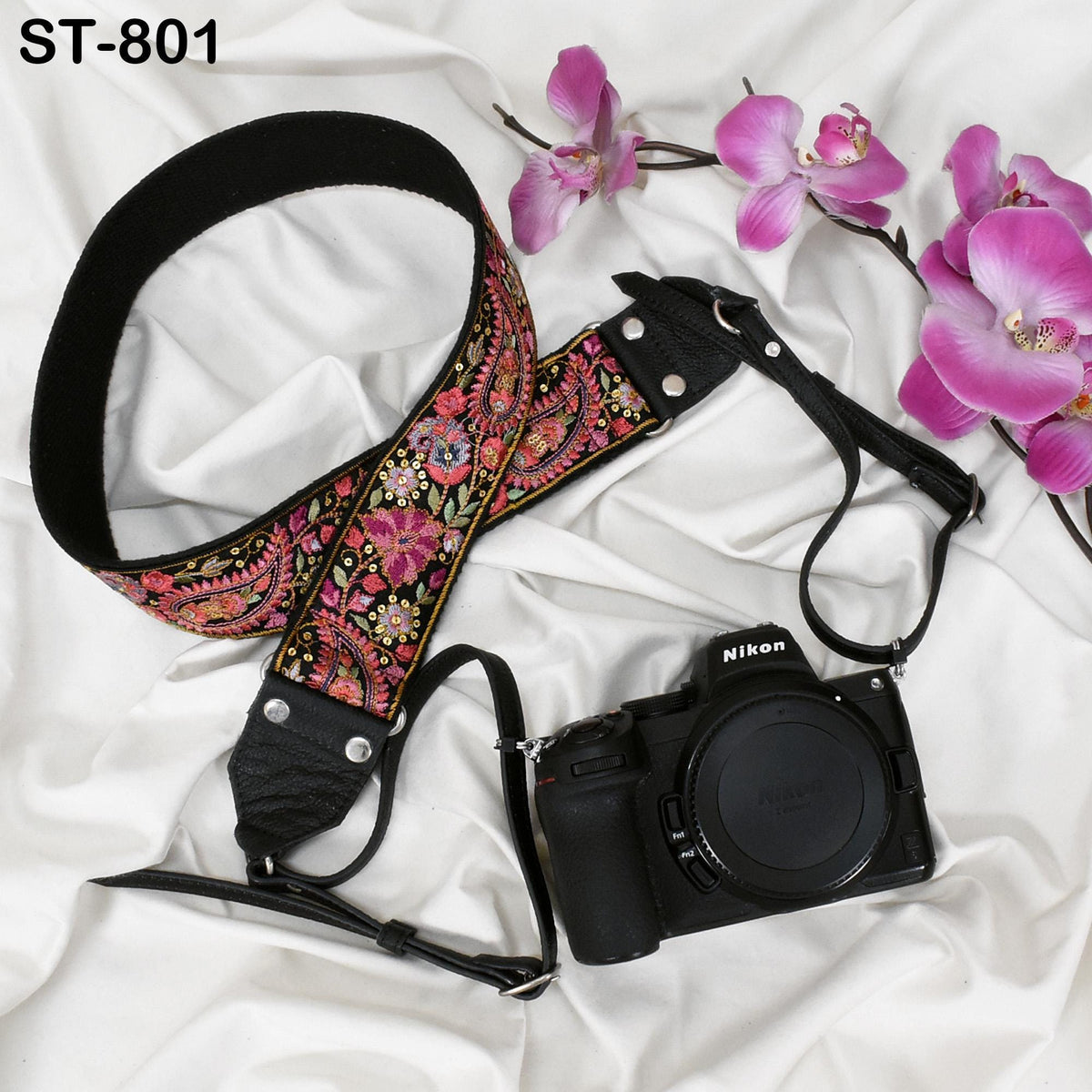 A black camera strap with a colorful, patterned design and a black camera body are placed on a white fabric surface, accompanied by pink orchids.
