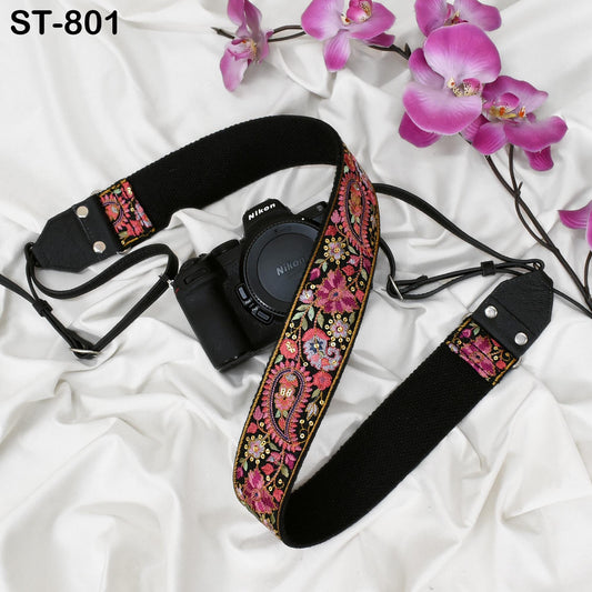 A black camera strap with a floral pattern and a black camera body are placed on a white surface, accompanied by pink orchids.