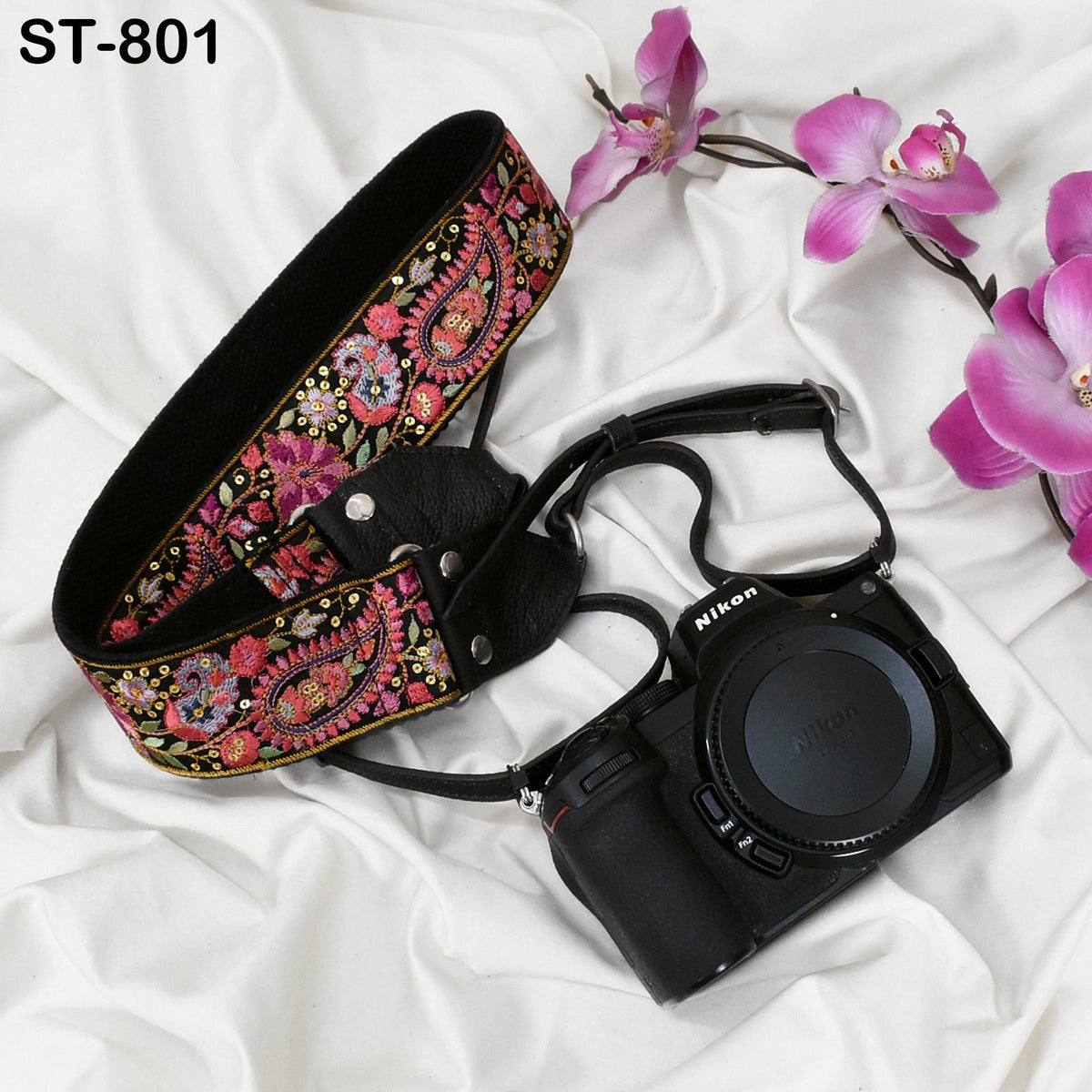 A floral patterned camera strap and a black Nikon camera are placed on a white fabric background, with pink orchids visible in the background.