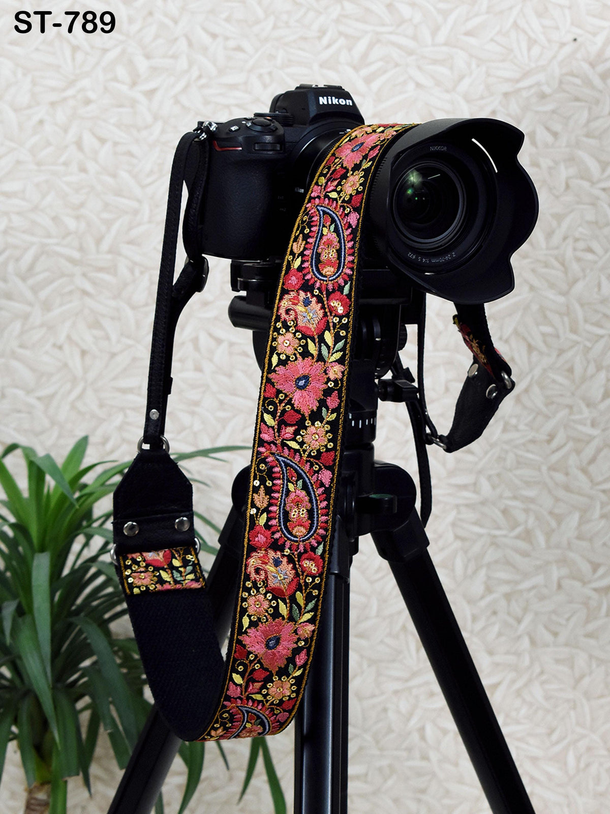 A black camera with a colorful floral patterned strap is mounted on a tripod, with a potted plant visible in the background.