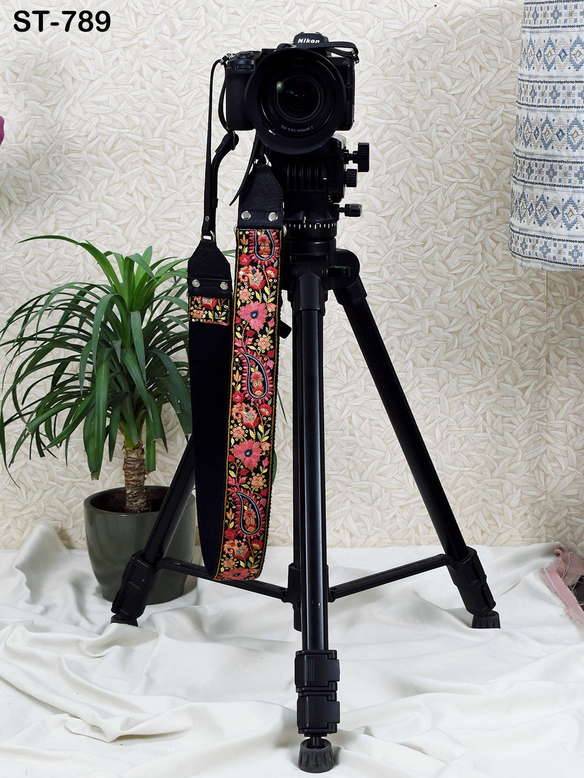 A black tripod with a camera mounted on top stands on a white cloth, accompanied by a potted plant in the background.