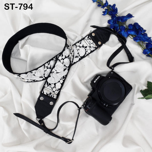 A black and white floral camera strap with a black camera attached to it, along with some blue flowers, all placed on a white cloth.
