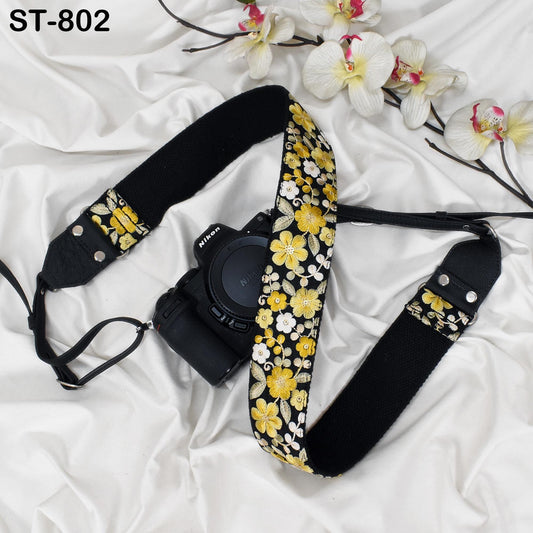 A black camera strap with a floral pattern is placed next to a camera lens, with white orchids and a white fabric surface in the background.