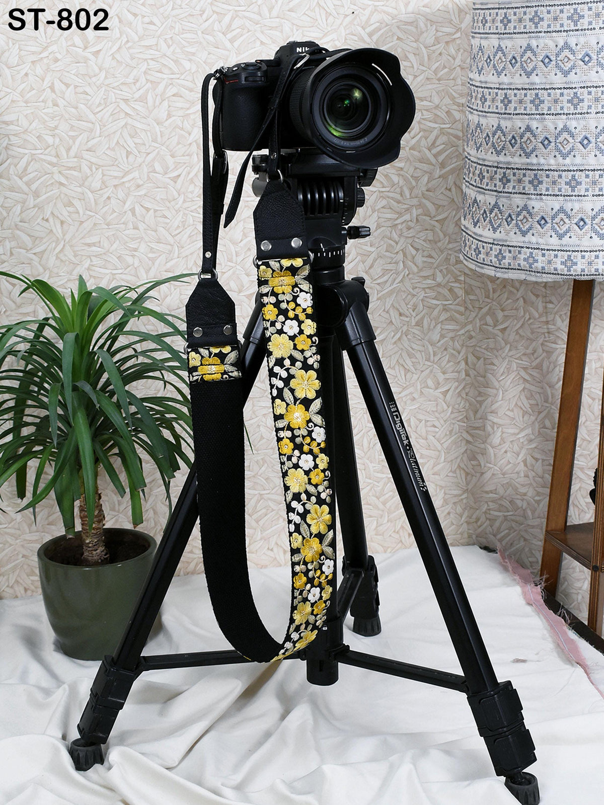 A black tripod with a camera attached to it, placed on a white surface next to a potted plant.