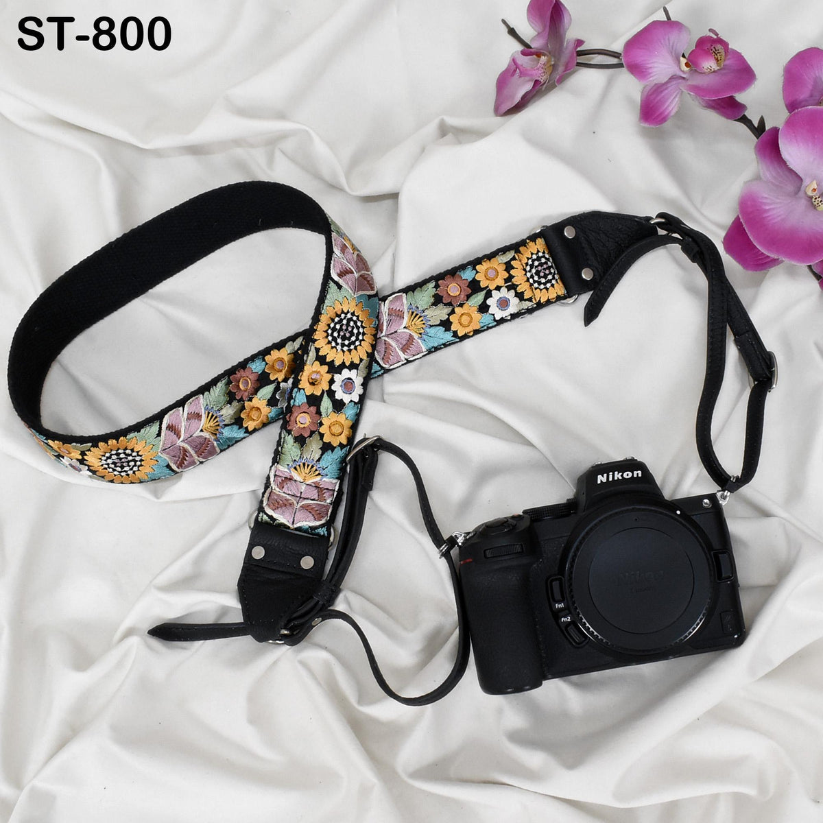 A floral patterned camera strap and a black camera with a lens cap are placed on a white fabric surface, accompanied by pink orchids.