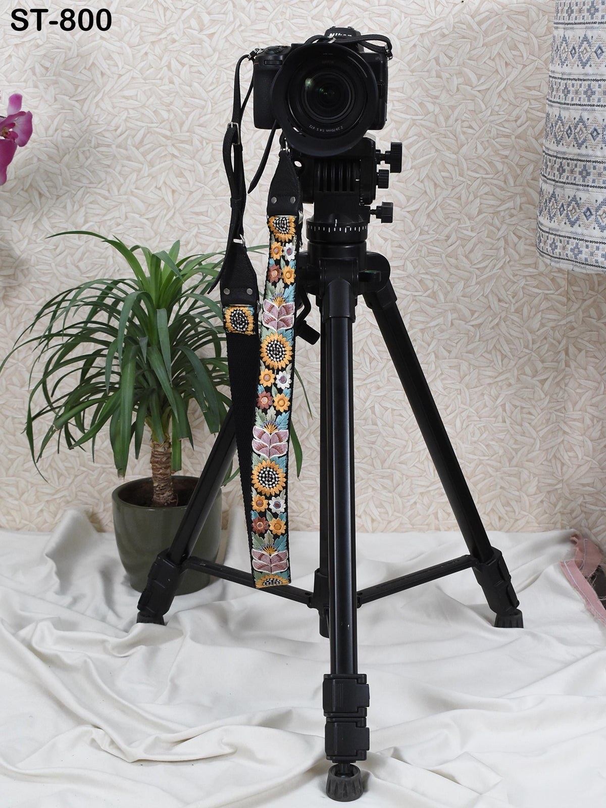 A black tripod with a camera attached to it stands on a white cloth, with a potted plant nearby.
