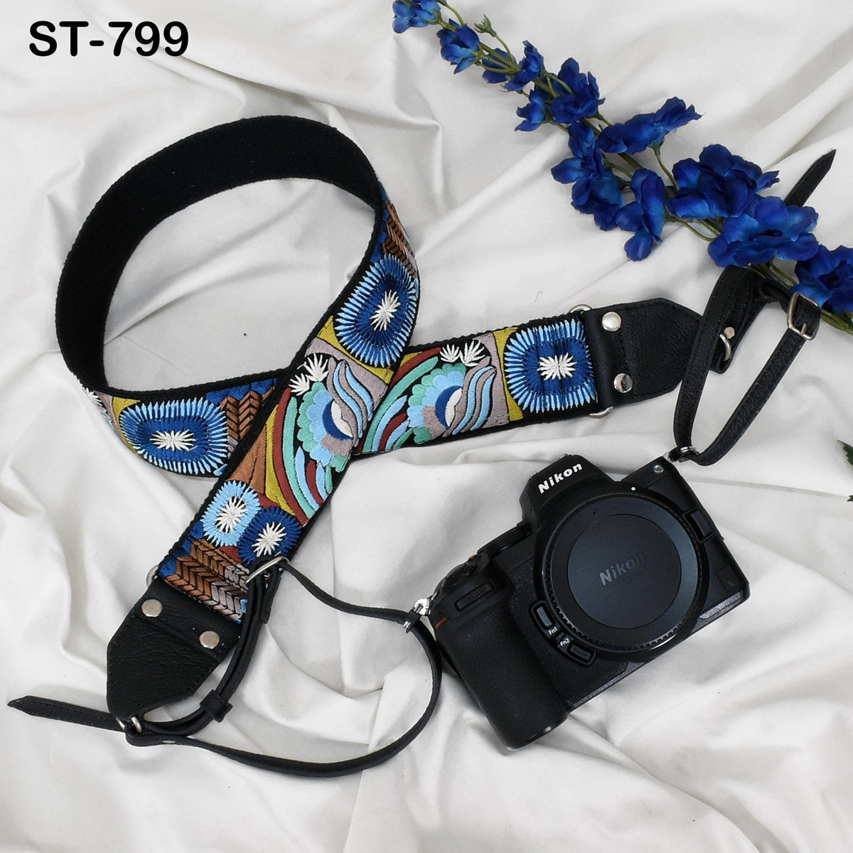 A colorful patterned camera strap and a black camera with a lens cap are placed on a white fabric surface, accompanied by a branch of blue flowers.