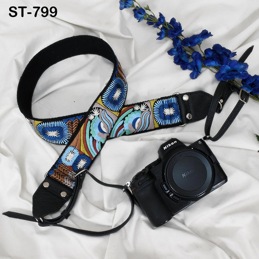 A colorful patterned camera strap and a black camera with a lens cap are placed on a white fabric surface, accompanied by a branch of blue flowers.