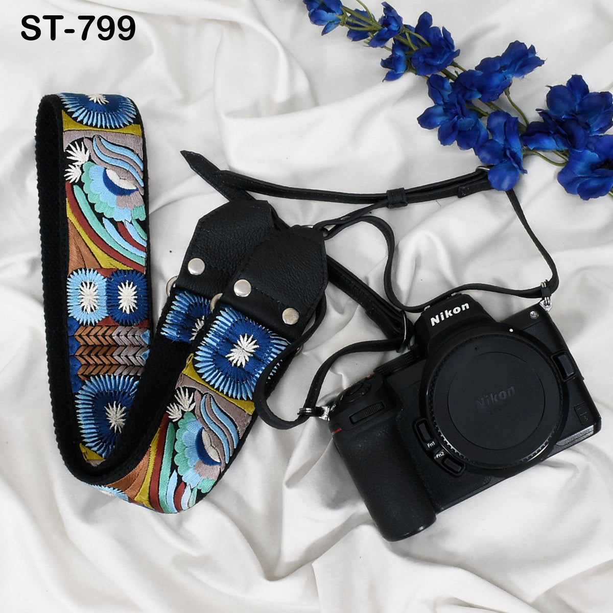A black camera with a colorful strap and a blue flower arrangement are displayed on a white surface.