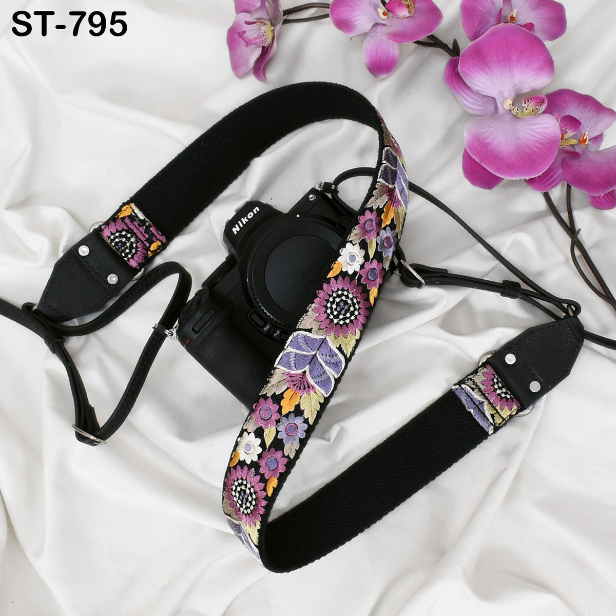 A black camera strap with a floral pattern is placed next to a pink orchid flower on a white background.