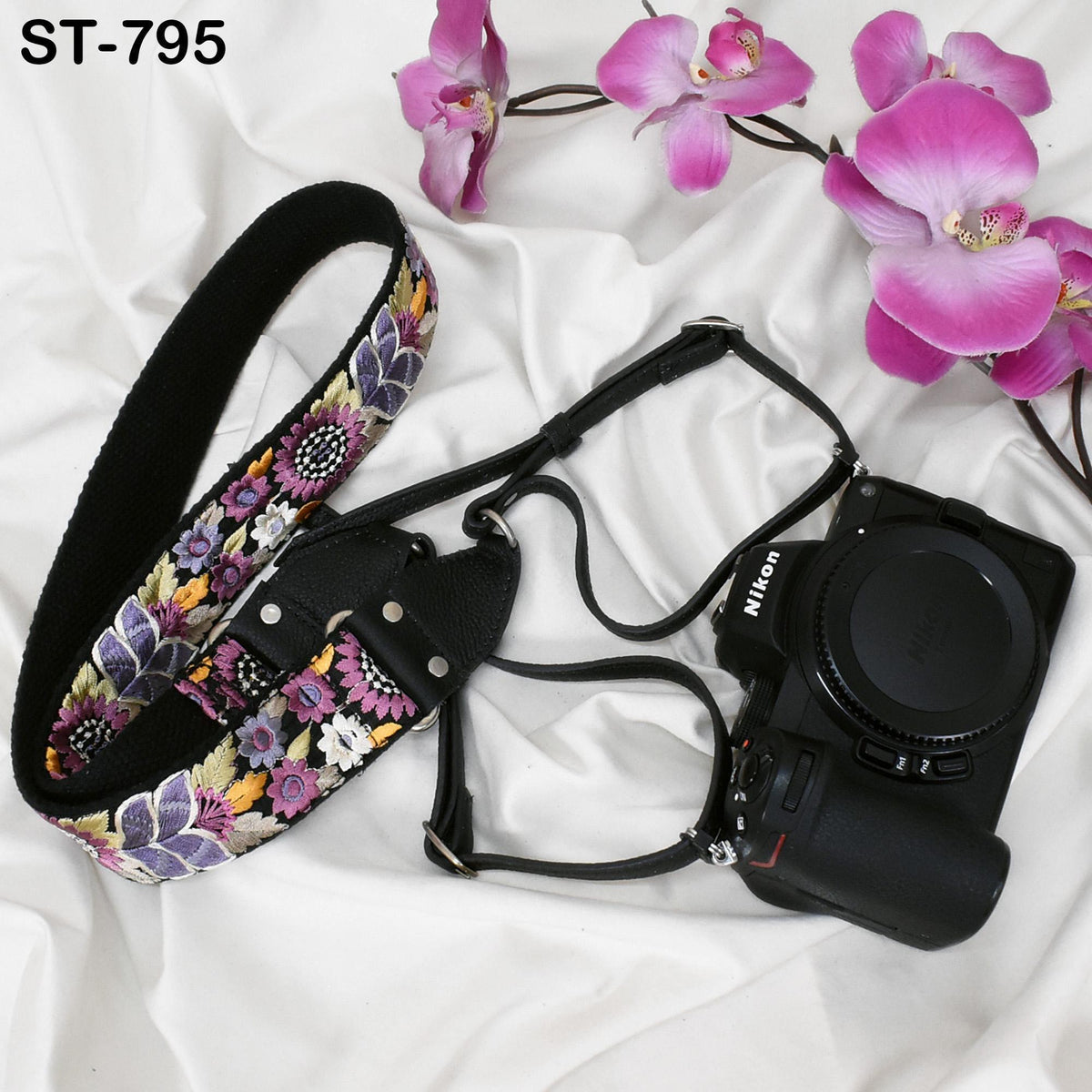 A floral patterned camera strap and a Nikon camera are placed on a white surface, with pink orchids in the background.