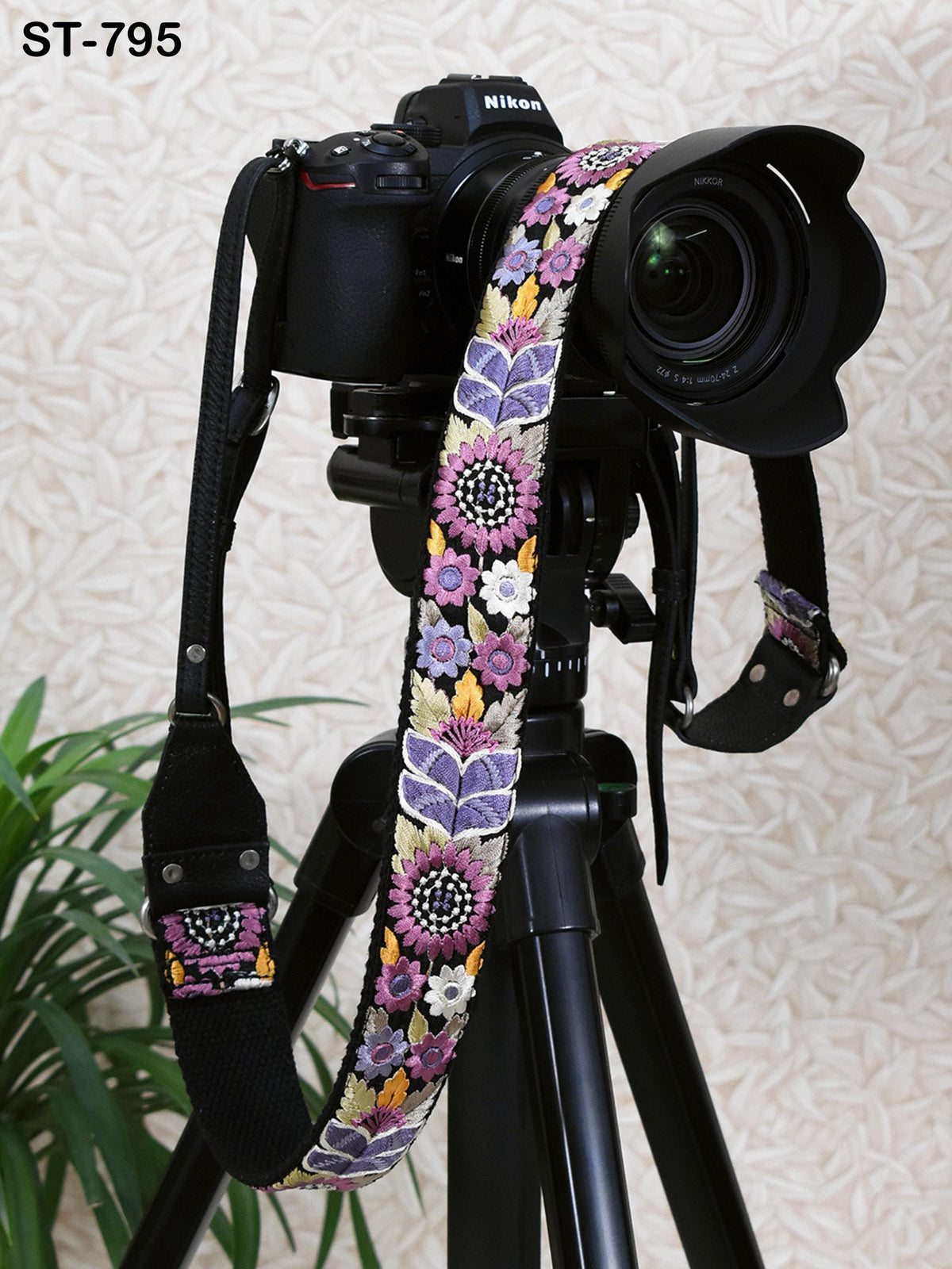 A black camera with a colorful floral patterned strap is mounted on a tripod, with a potted plant visible in the background.
