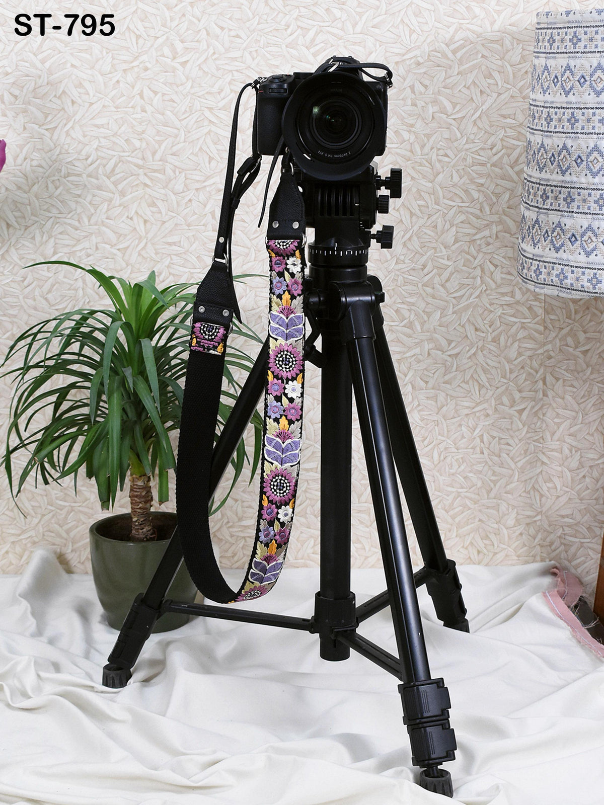 A black tripod with a camera mounted on top stands on a white cloth, accompanied by a potted plant in the background.