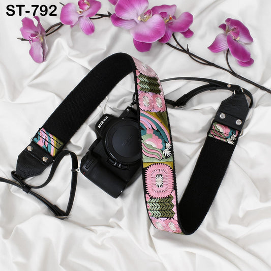 A black camera strap with a colorful floral pattern is placed next to a pink orchid flower on a white surface.