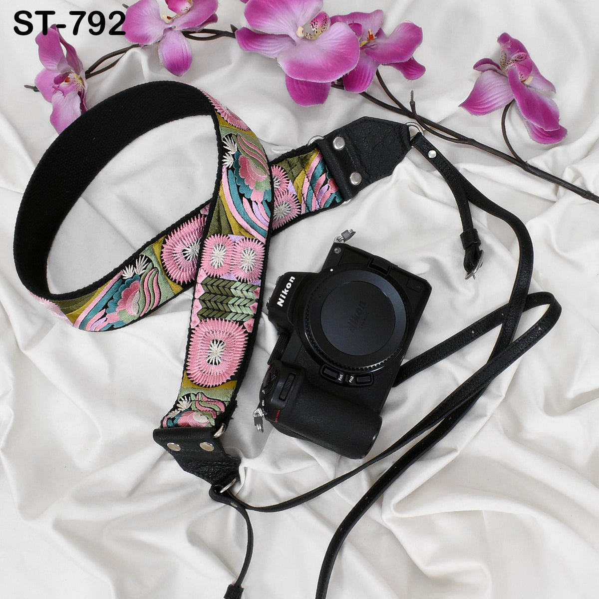 A floral patterned camera strap and a black camera with a lens cap are placed on a white surface, accompanied by pink orchids.