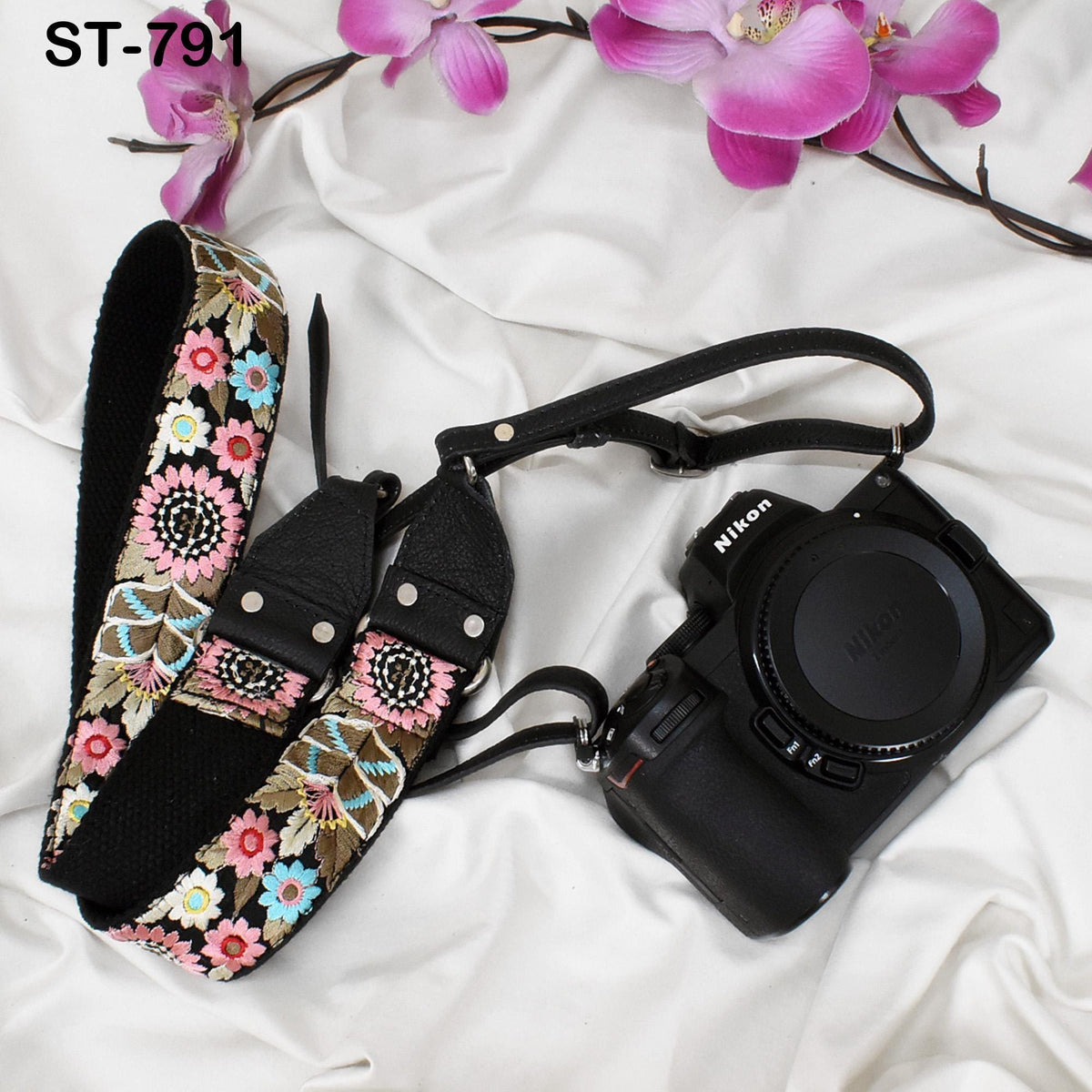 A floral patterned camera strap and a black Nikon camera are placed on a white surface, with pink orchids in the background.