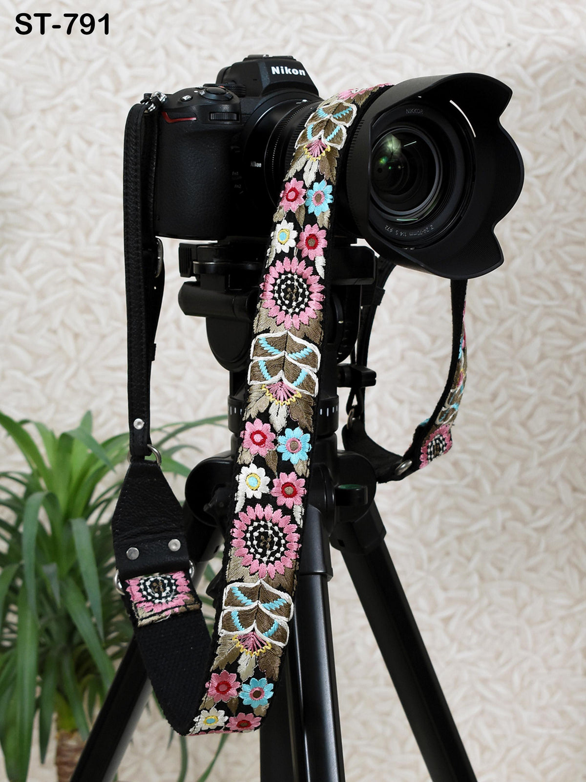 A black camera with a colorful floral strap attached to it, placed on a tripod against a patterned wall.