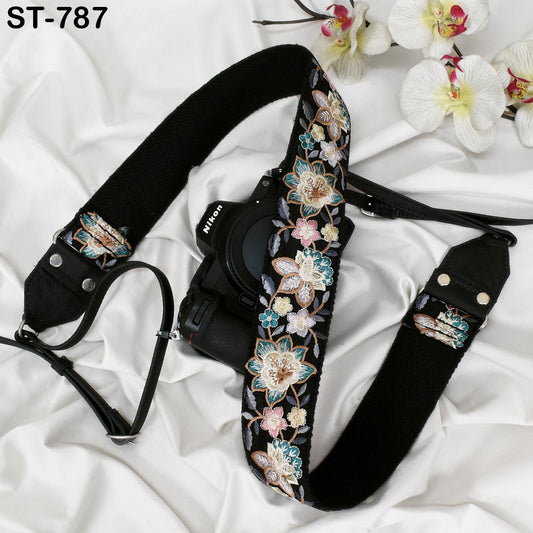 A black camera strap with a floral pattern is placed on a white surface, accompanied by a few white orchids.