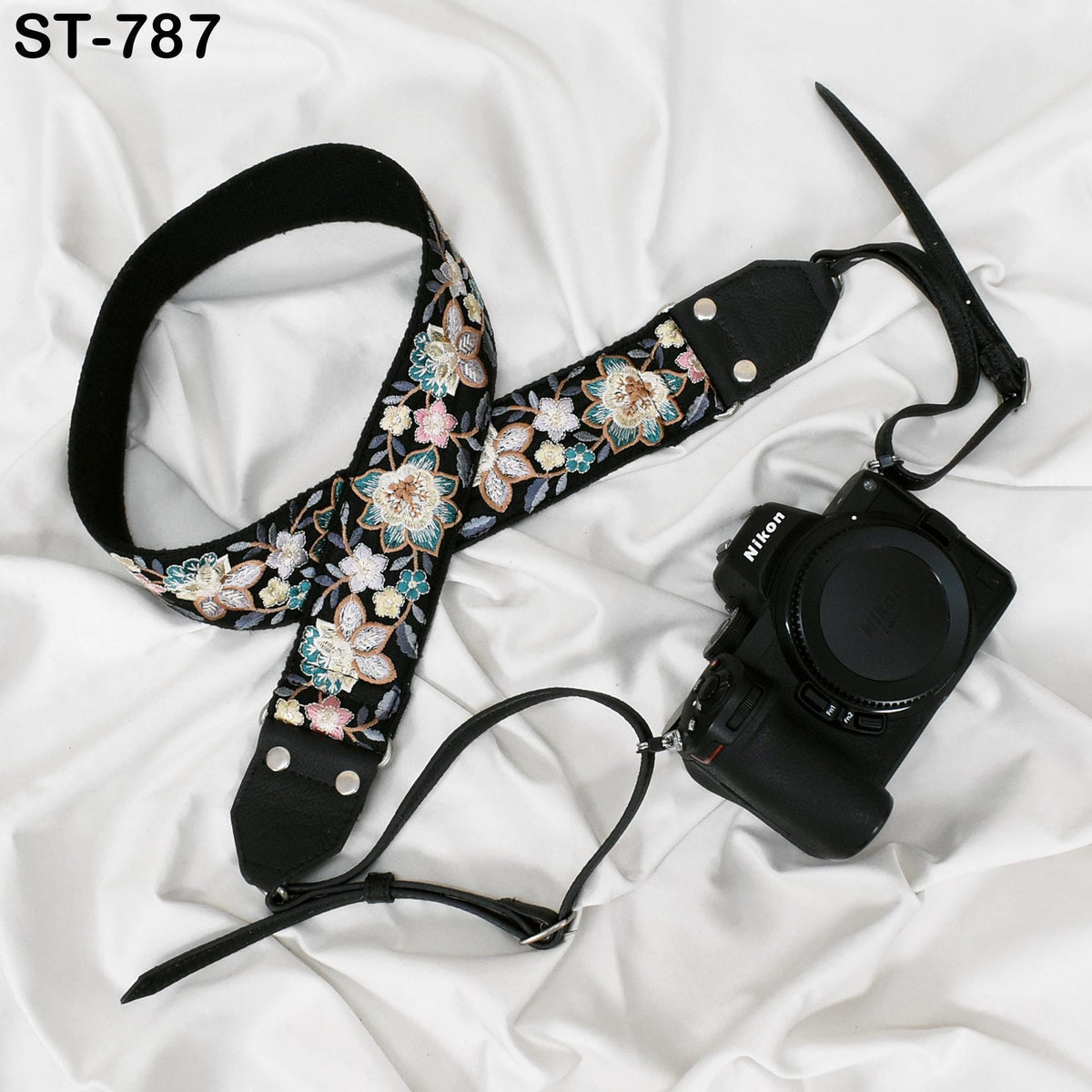 A floral patterned camera strap and a black camera with a lens attached are placed on a white fabric surface.
