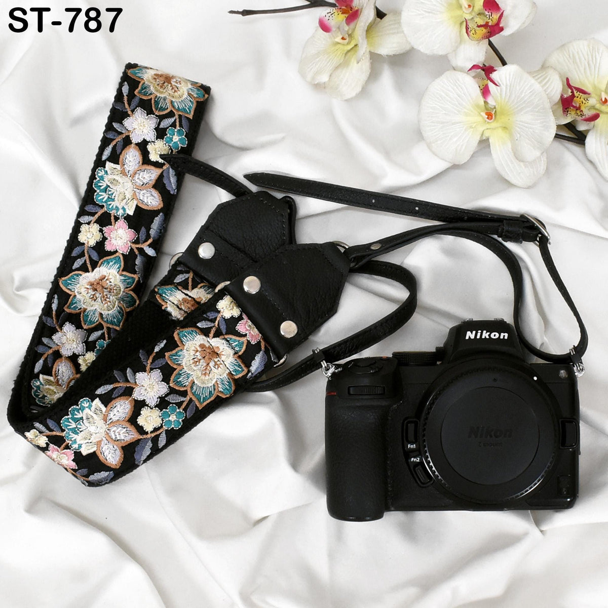 A floral patterned camera strap and a Nikon camera are placed on a white background, with white orchids in the background.