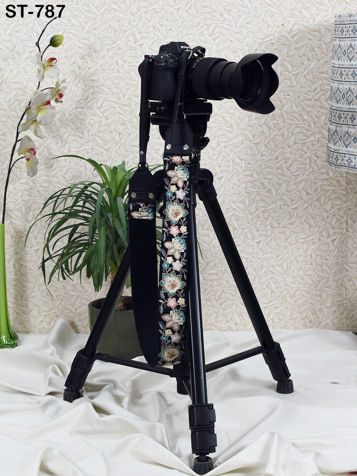 A black camera tripod with a camera attached to it stands on a white surface, with a potted plant and a vase nearby.
