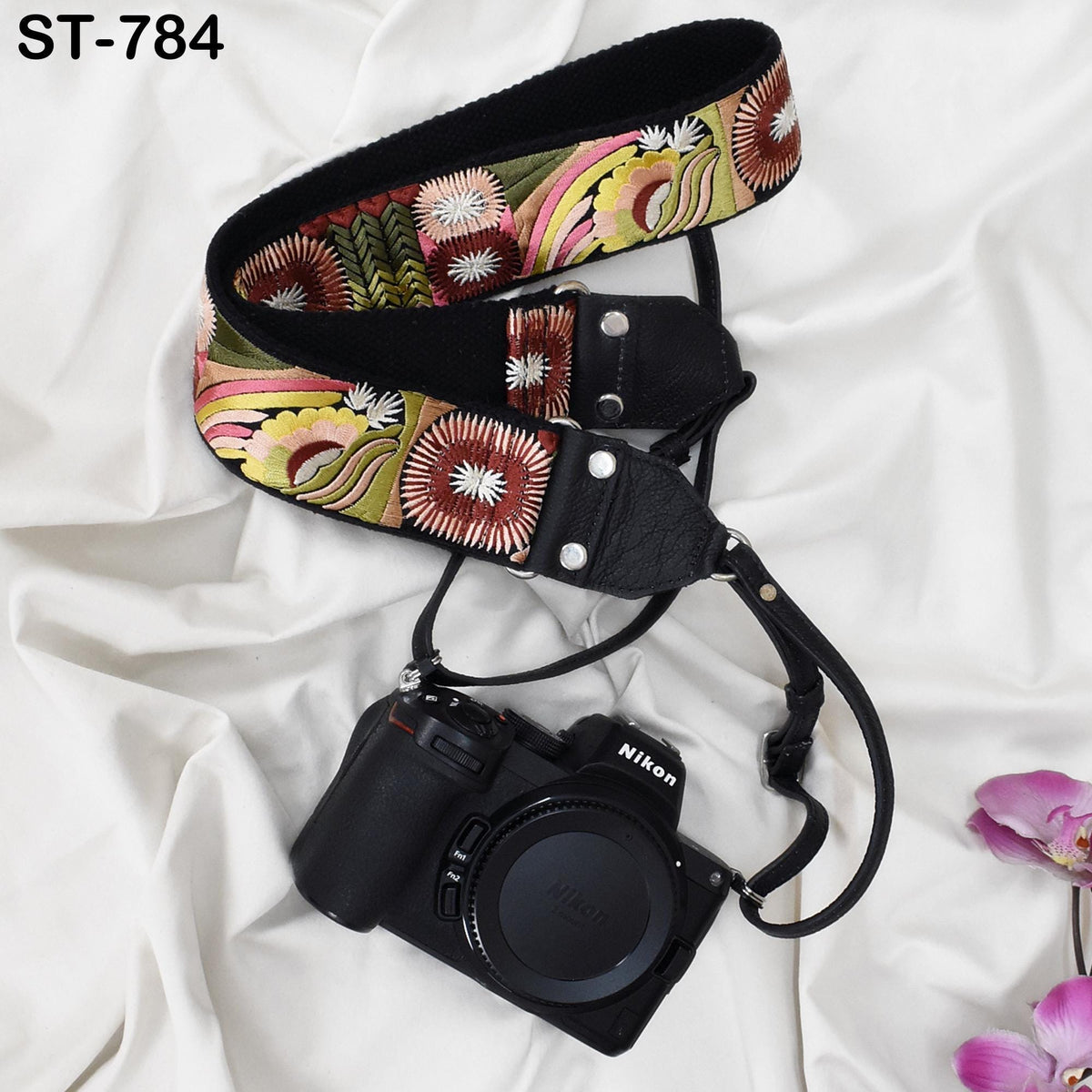 A black camera with a colorful floral patterned strap is placed on a white fabric surface, accompanied by a few purple flowers.