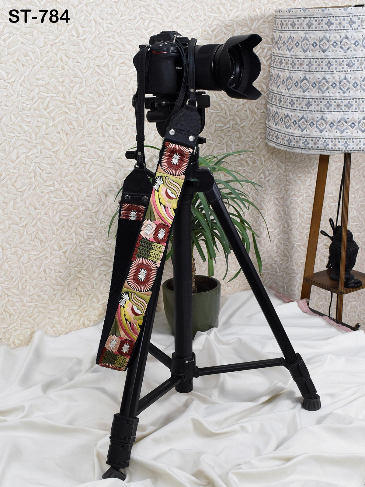 A black tripod with a camera attached to it, along with a colorful strap, is placed on a white surface. There is a lamp and a plant in the background.