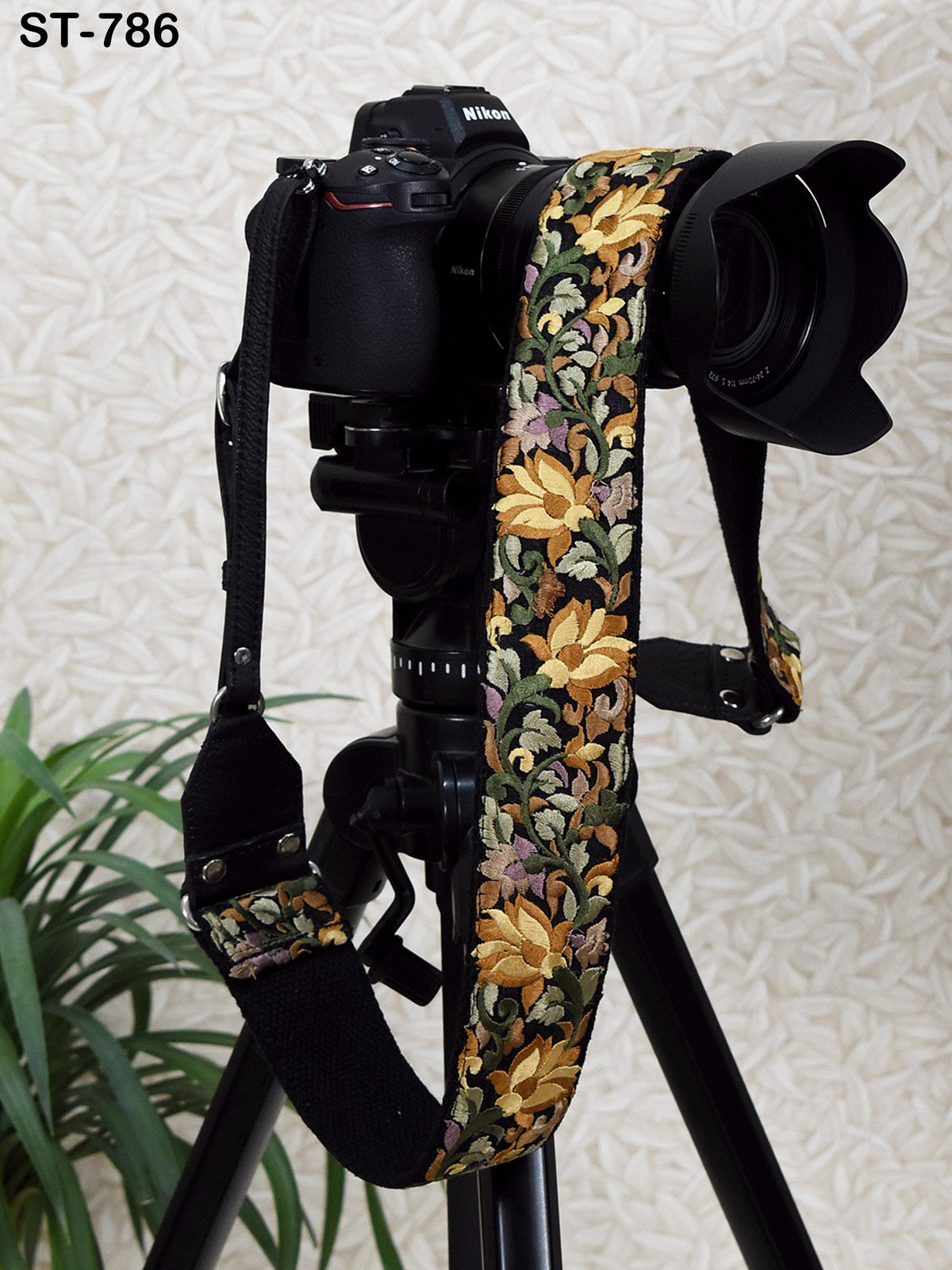 A black camera with a floral patterned strap is mounted on a tripod, with a potted plant visible in the background.