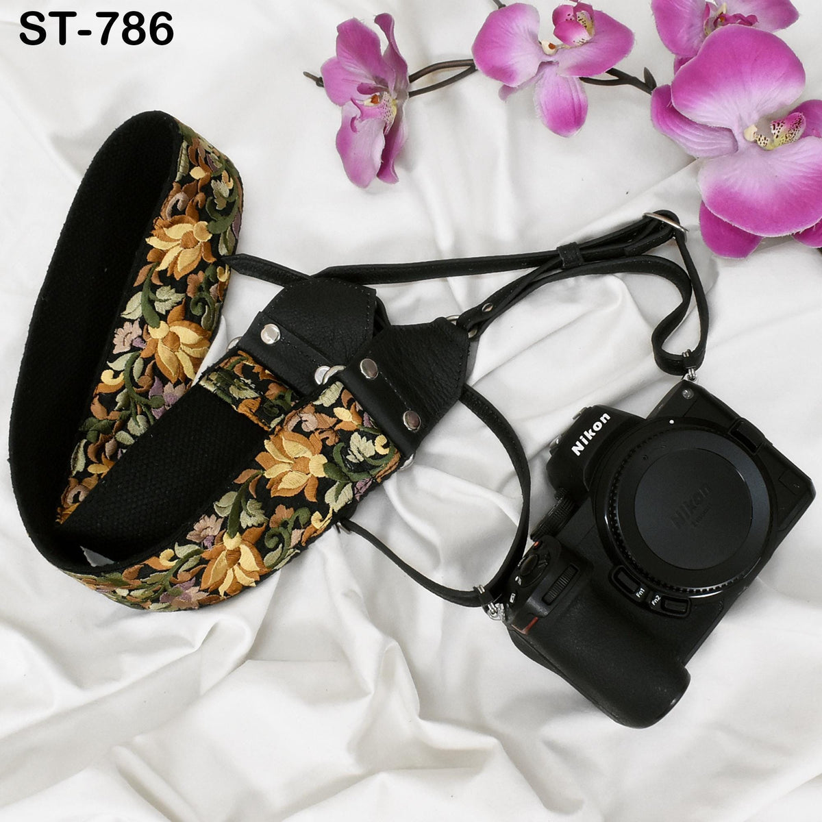 A floral patterned camera strap and a Nikon camera are placed on a white surface, with pink orchids in the background.