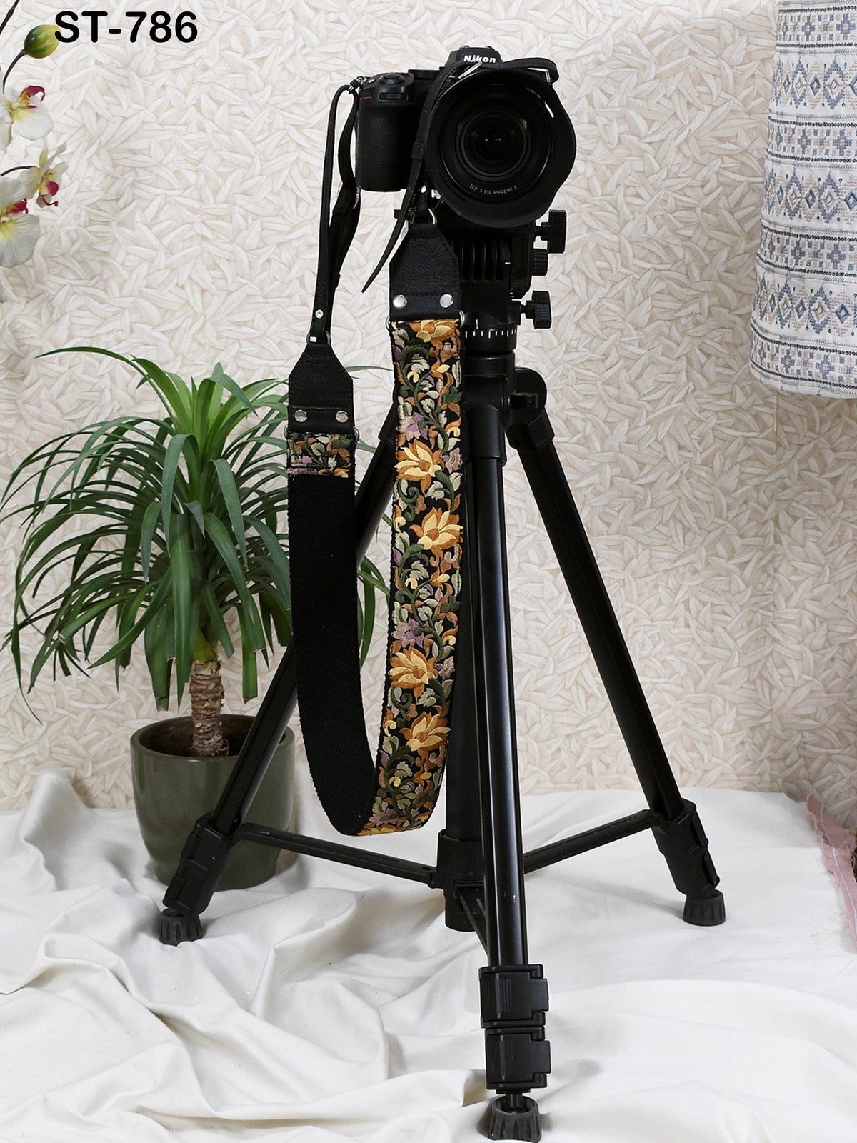 A black tripod with a camera attached to it, placed on a white surface next to a potted plant.