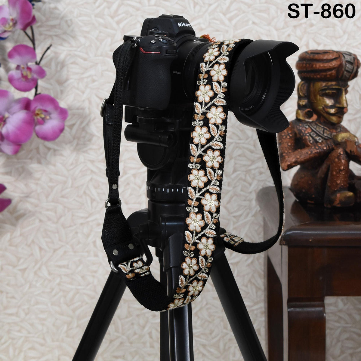 A black camera with a floral patterned strap is mounted on a tripod, with a statue of a seated figure visible in the background.