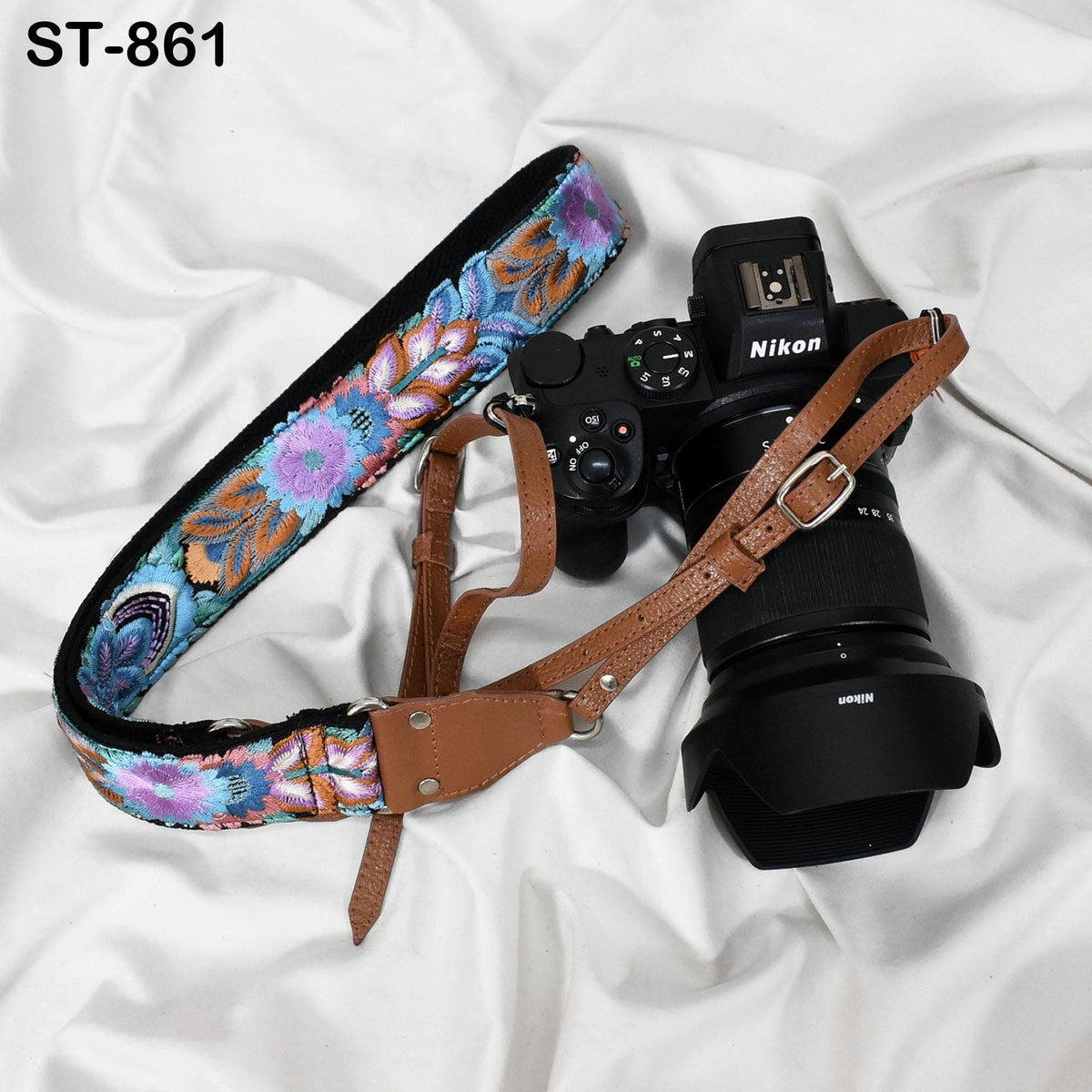 A floral-patterned camera strap and a Nikon camera body are placed on a white fabric surface.