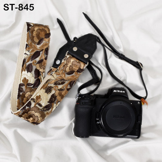 A black and brown floral camera strap and a black Nikon camera are placed on a white surface.