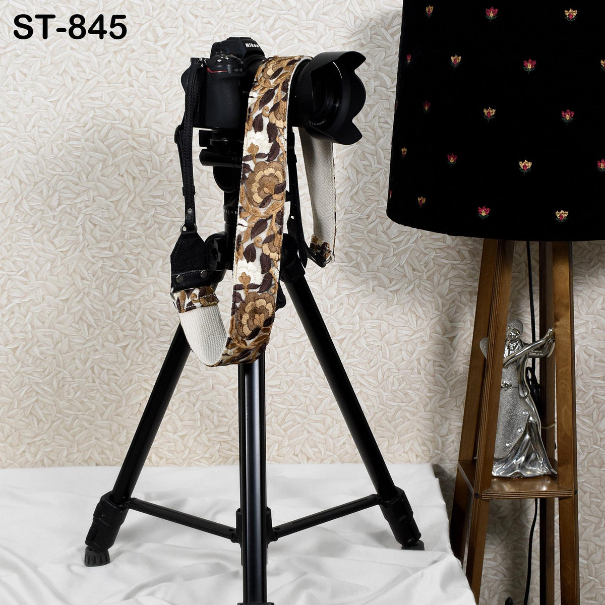 A black tripod with a camera attached to it, standing on a white surface next to a black lamp with a floral pattern.