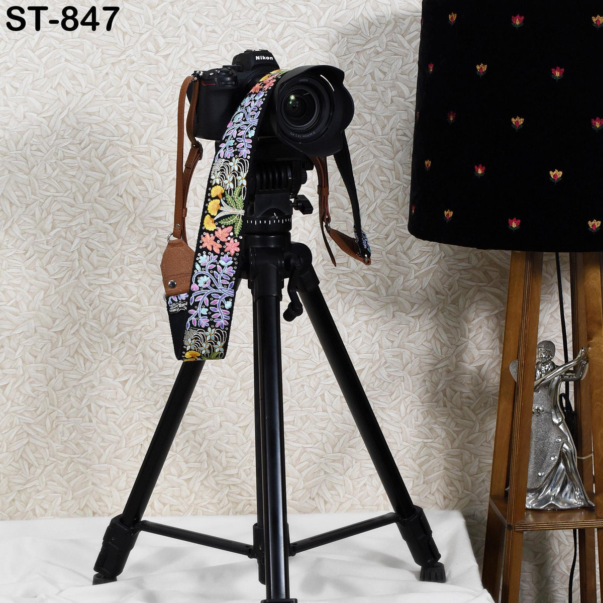 A black tripod with a camera attached to it, placed on a white surface next to a lamp.