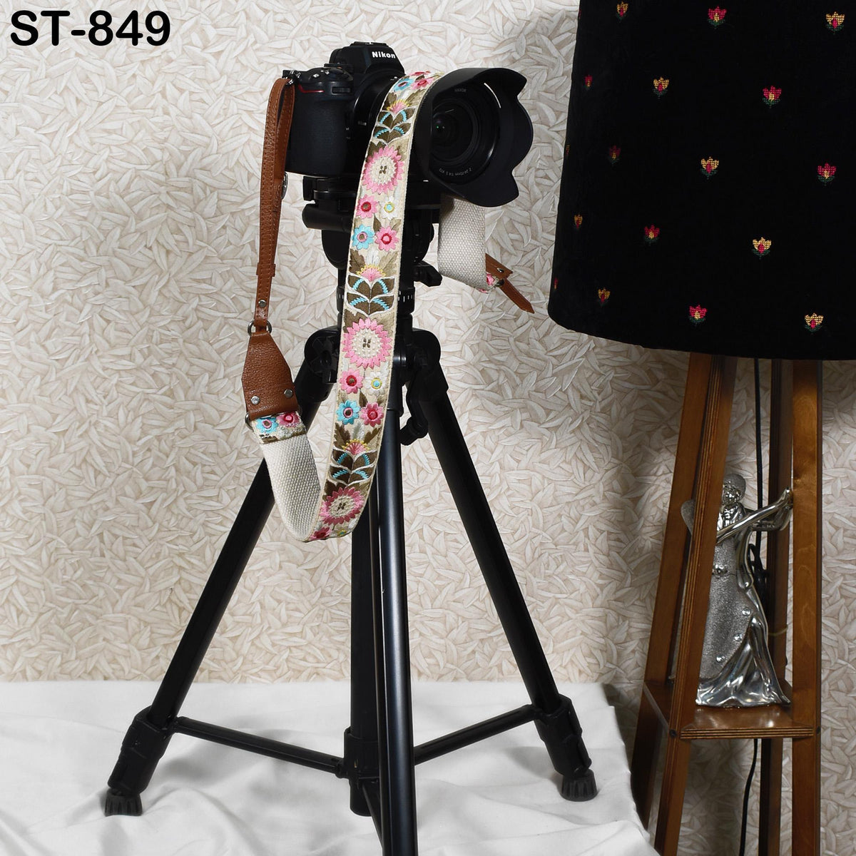 A camera mounted on a tripod, with a floral patterned strap, is placed next to a lamp on a wooden stand.