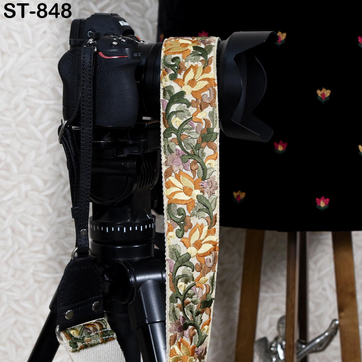 A black camera with a colorful floral patterned strap is mounted on a tripod, with a black background and a wooden stool visible in the background.