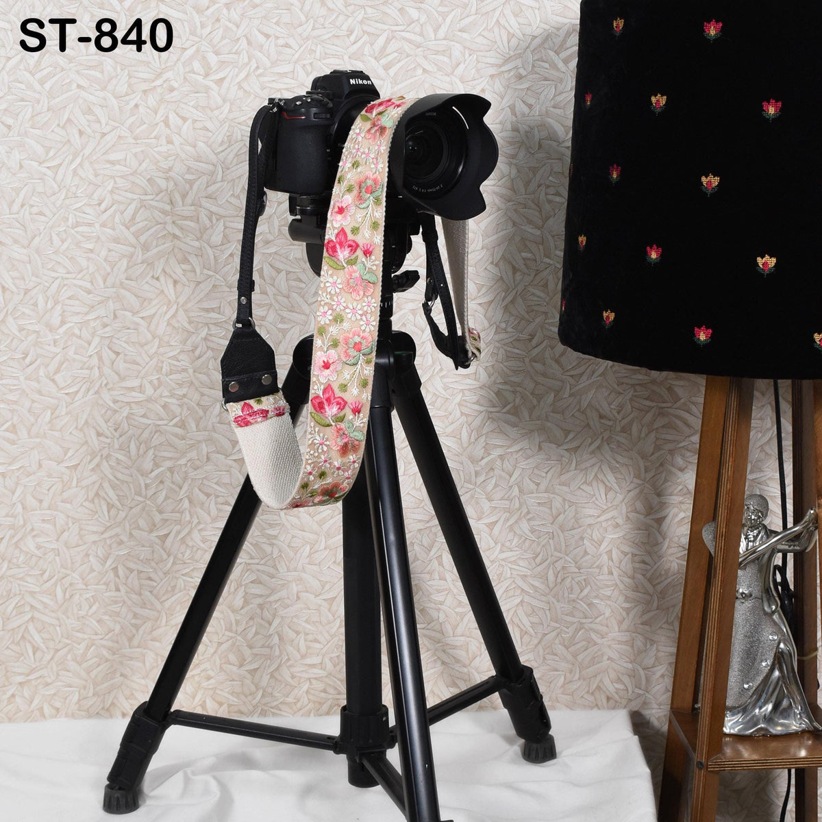 A black tripod with a camera attached to it, along with a floral patterned strap, is placed next to a lamp.