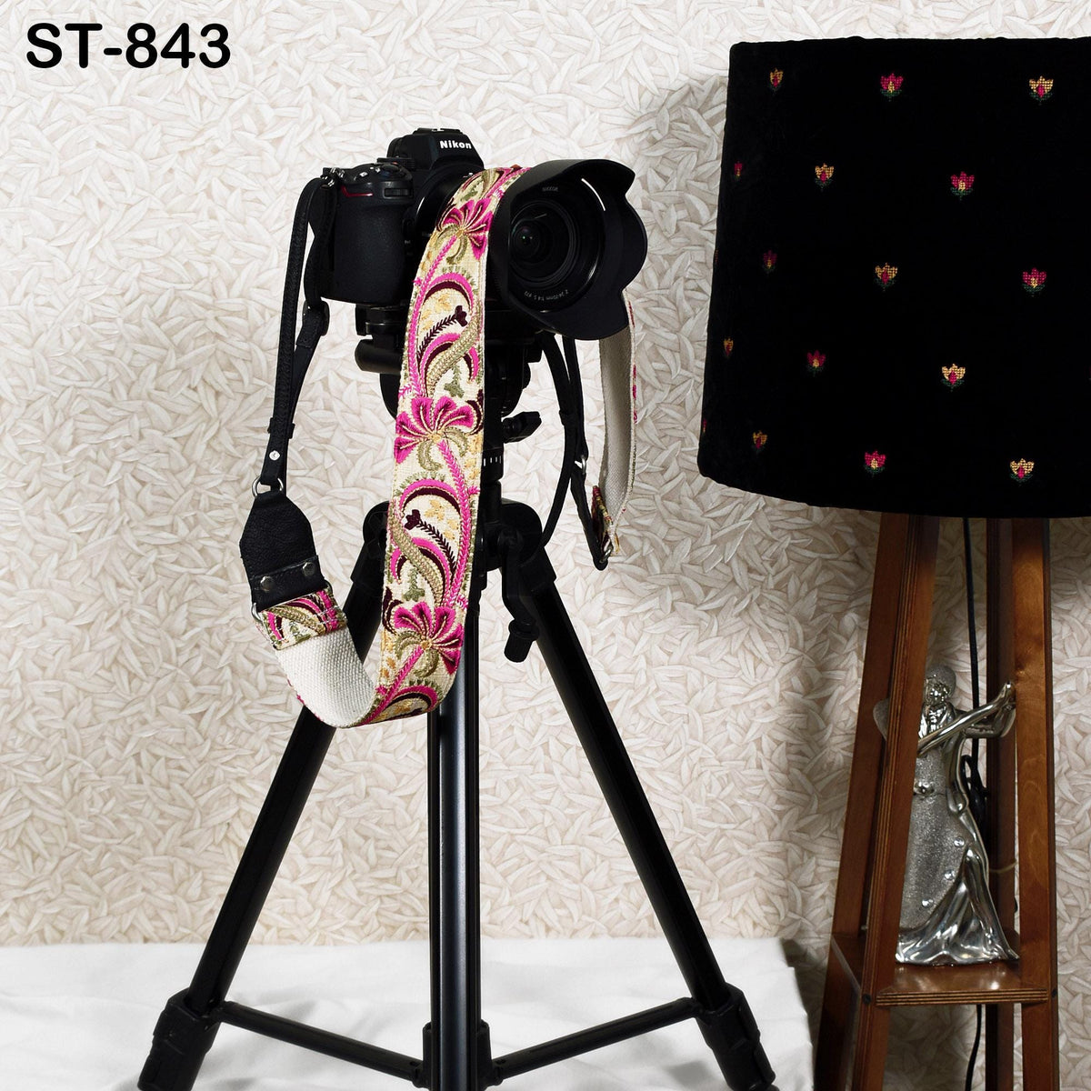 A black tripod with a camera mounted on top, along with a decorative floral strap, stands next to a black lamp with a patterned lampshade.