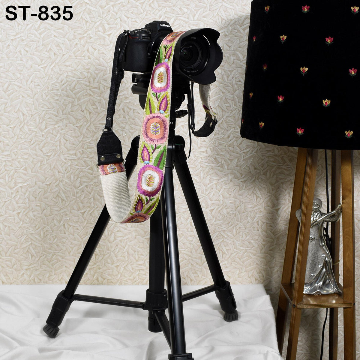 A camera mounted on a tripod, with a colorful strap attached, is placed next to a lamp on a wooden stand.