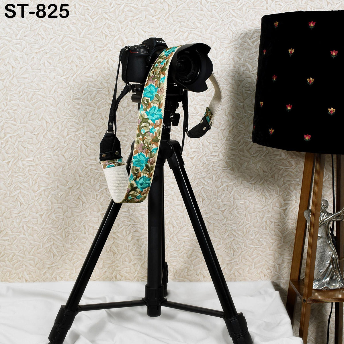 A black tripod with a camera mounted on top, along with a floral patterned strap, stands on a white surface next to a black lamp.