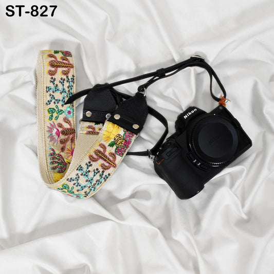 A floral patterned camera strap and a black camera are placed on a white fabric surface.