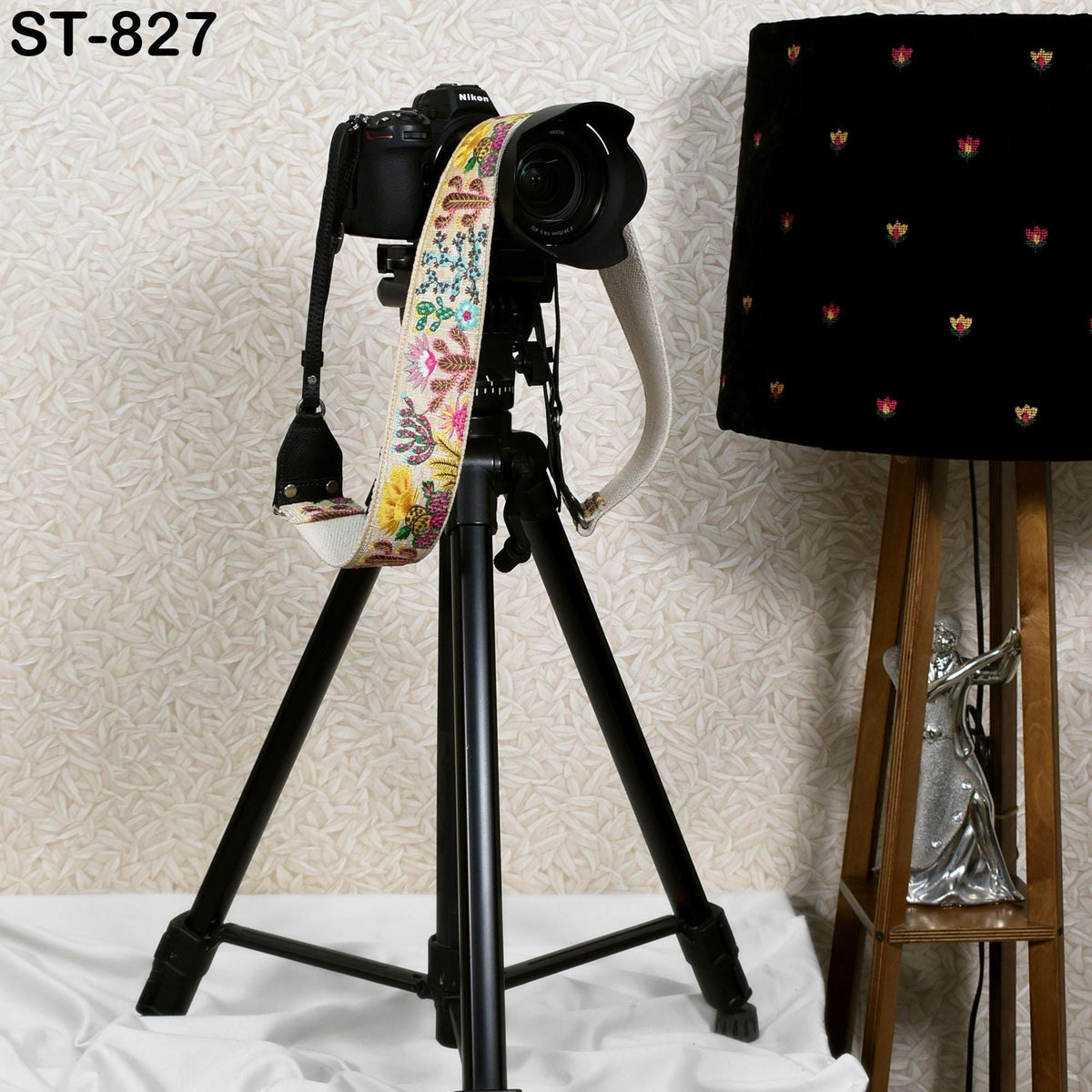 A camera mounted on a tripod, with a colorful strap attached, is placed next to a lamp on a wooden stand.