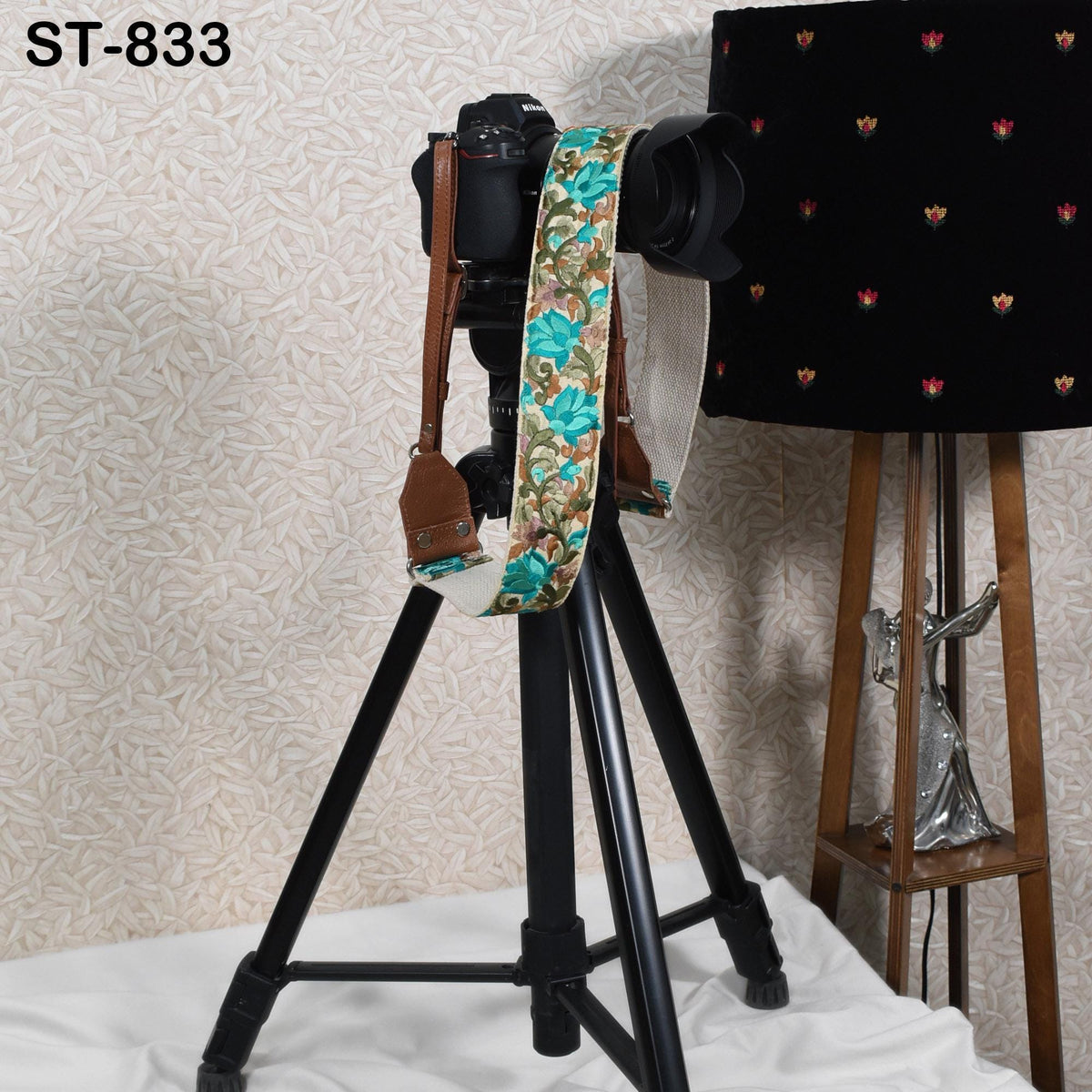 A black tripod with a camera strap attached to it, placed on a white surface next to a black lamp with a floral pattern.