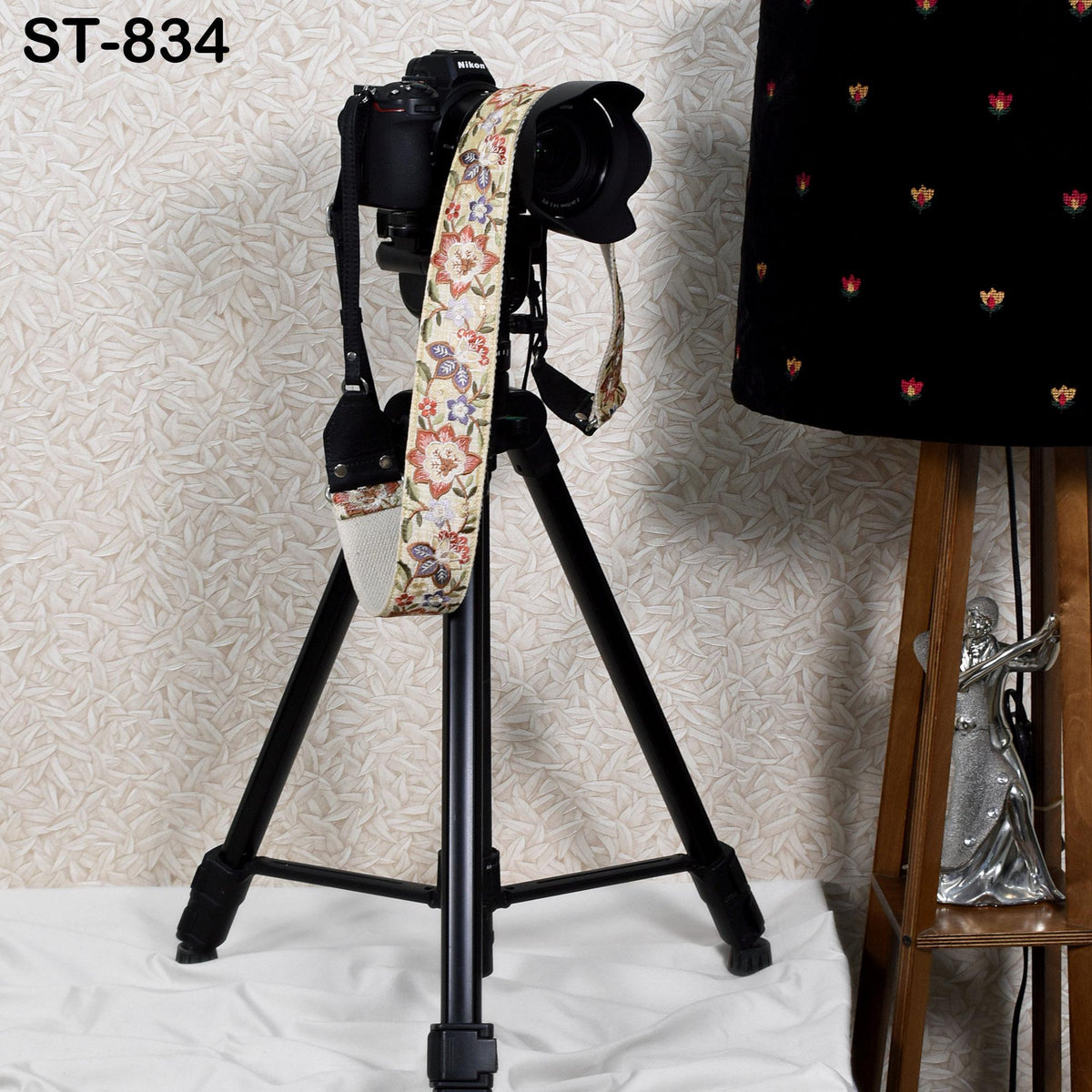 A black tripod with a camera strap attached to it, standing on a white surface next to a black lamp with a floral pattern.