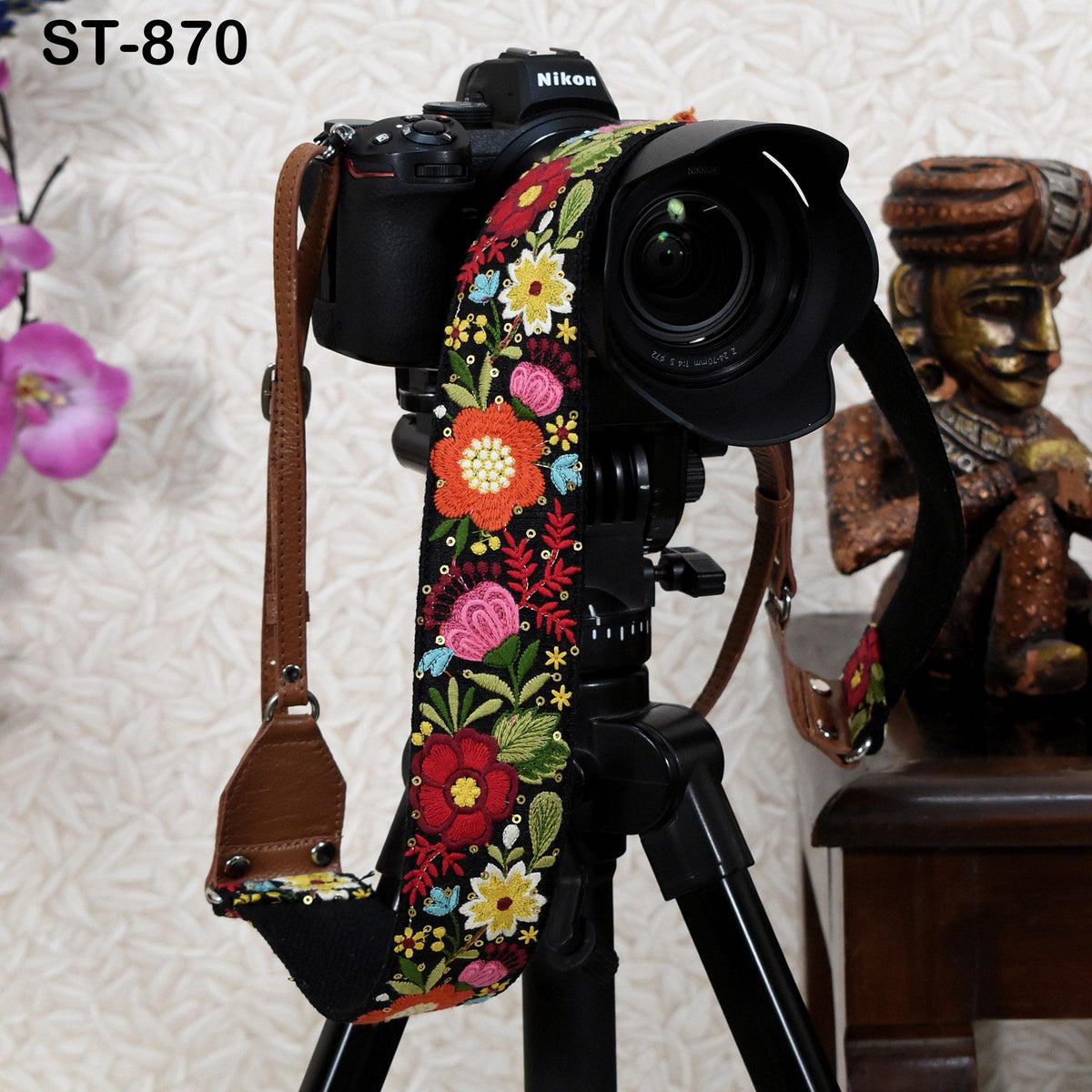 A black camera with a colorful floral strap is mounted on a tripod, with a wooden statue visible in the background.